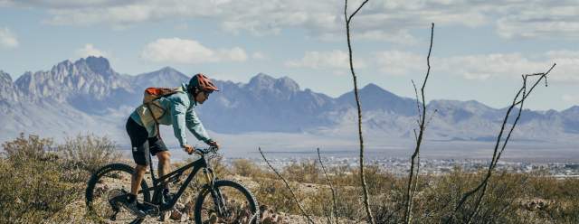 Person riding a mountain bike on a desert trail with rugged mountains in the background.