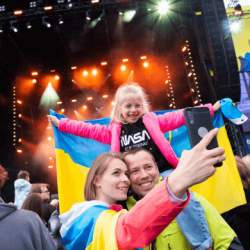 A young girl sits on a mans shoulder, holding a Ukrainian flag. The family are taking a selfie