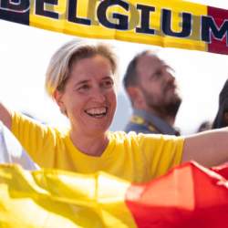 A lady in a yellow tshirt smiles holding a flag with the words 'belgium' on there