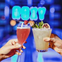 Two people 'cheers'ing two cocktails in front of a neon sign saying 'ROXY'.