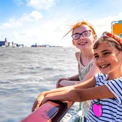 Two young girls stand on the Mersey Ferry smiling on a sunny day
