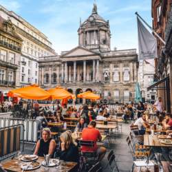 A bustling street in Liverpool with people sitting at outside tables with umbrellas and people eating