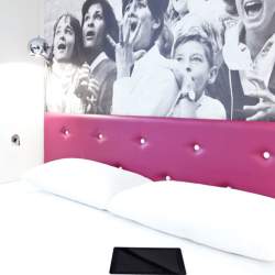 A double bed in a hotel room. There are white bed sheets and a pink, leather-look headboard. Behind the bed is a black and white photo of an excited crowd.