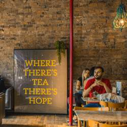 Inside the LEAF cafe. Against an exposed brick wall is a large print that reads 'where there's tea there's hope.' People are sitting at wooden tables and chairs in various chalky colours. There's also a disco ball hanging from the ceiling and a large red supporting column.