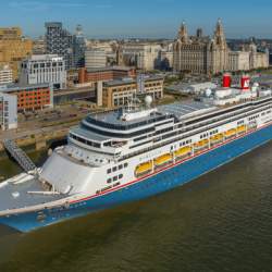 A cruise ship docked on Liverpool Waterfront. The bottom of the ship is painted blue, there are yellow hanging life boats and red chimneys.