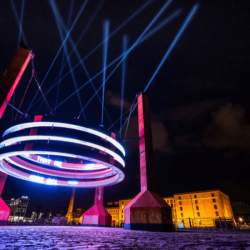A huge structure of silver rings suspended on four large, red ballasts. Laser lights are shining into the sky in various directions at night.