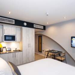 Inside an aparthotel that is airy and bright with a large white bed, a small kitchenette and flat screen TV on a whitewall.