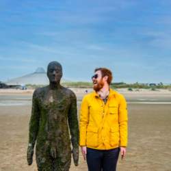 A man in a yellow rain coat stands next to a metal statue on Crosby Beach