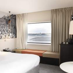 A light, spacious hotel room with a large bed with white sheets. Behind the bed is a black and white historic image of Liverpool Docklands. There's a cushioned bench style seat underneath a window, with beige curtains open.