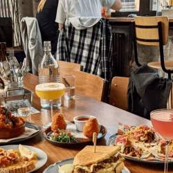 A selection of food on small plates. The food is served with two bright cocktails in martini style glasses. The room is light and airy and has bench seating.