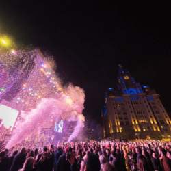 A crowd of people in front of the main stage on the Liverpool Pier Head at night time. The Royal Liver Building can be seen lit in blue in the night sky.
