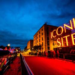 A large red neon sign that reads 'ICONIC SITE' alongside the Royal Albert Dock warehouses