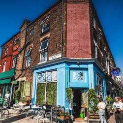An exterior shot of a Victorian Style corner building with small sash windows and pastel blue shop front below. There are lots of plants decorating the shop front with tables and chairs outside on a sunny day.