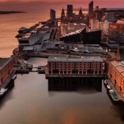 A drone shot over Liverpool looking across Royal Albert Dock warehouse buildings and the Pier Head with orange sky