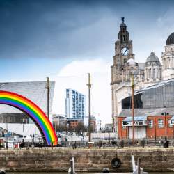 A cross section image looking across towards Liverpool Pier Head. It is an Autumn day with a greyish, cloudy sky. A giant rainbow ads colour alongside the modern Museum of Liverpool building which is contrasted by the Royal Liver Building and Port of Liverpool Building.