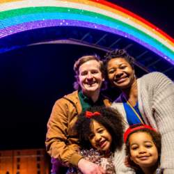 A family smiling in front of a giant 50ft rainbow lit up