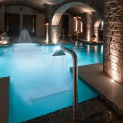 A spa pool inside a vaulted, brick space with low lighting and lanterns. The pool is blue and has hydro taps pouring into the pool.