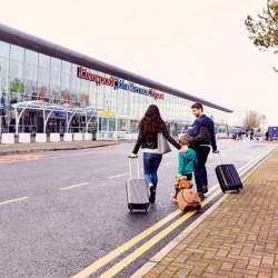 A man and woman are pulling small suitcase. There is a young child between them pulling a teddy bear suitcase. They are outside a modern, glass fronted building which is Liverpool Airport