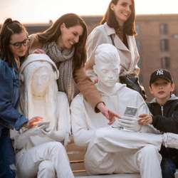 Two adults and two children engage with a statue of people painted white, sitting on a bench looking at 'smart phones.'