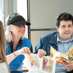 A man in a denim jacket reads a story, while a woman in a visor on her head next to him shows the pages of the book to the audience of children