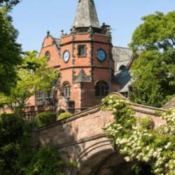 A red brick bridge covered in greenery. Behind is a large red brick church with a turret roof and clock face.