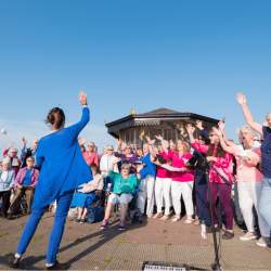 A choir singing on New Brighton promenade. The choir leader is wearing a bright blue outfit and the members of the choir have their hands in the air as they sing.