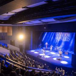A stage with blue lighting and people on stage performing, there is a large crowd watching the performance too.