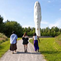 3 people walking in a grassy area surrounded by trees looking on to a large whites sculpture of a young girls head