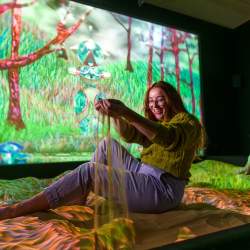 A lady sits in a sandpit in a dark room with a large, projected screen of nature behind her