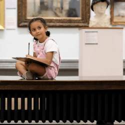 A young girl sitting on a bench and drawing, whilst in a museum.