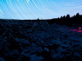 Long-exposure night sky with star trails over a snow-covered landscape, with a person holding a red light near a tree line