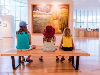 Three kids sitting on a wood bench in front of a landscape painting at the Southern Utah Museum of Art.