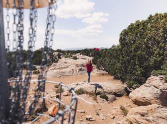 A person dressed in red throwing a disc into a disc golf basket in Three Peaks Recreation Area