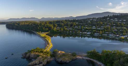 Aerial view of a narrow shoreline separating ocean and lagoon with forest and homes beyond.