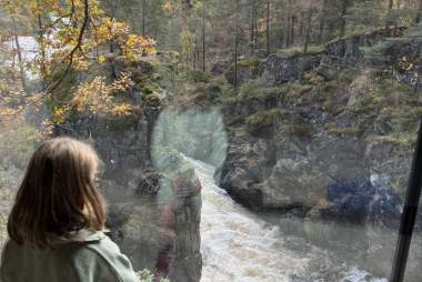 Et barn sitter ved et stort vindu og ser ut på Kvåsfossen i Lyngdal. Elva fosser ned gjennom kløften omgitt av høstfargede trær.