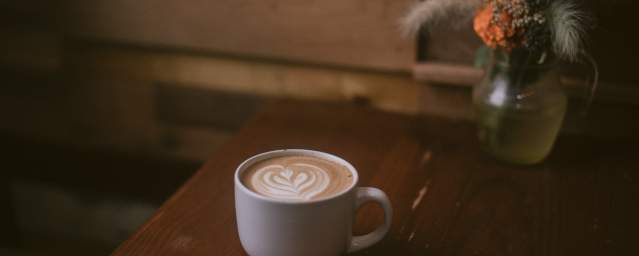 A latte served in a white cup sits on a table next to a vase with orange and yellow flowers.