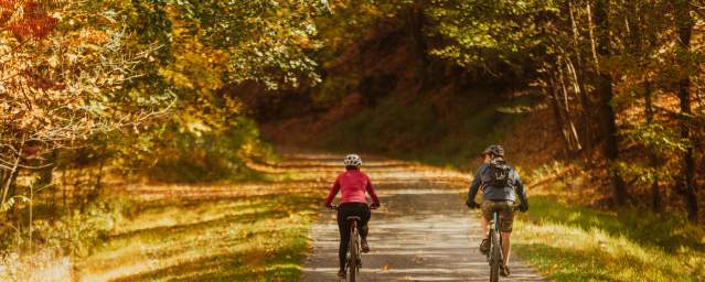 A couple ride bike on a gravel path surrounded trees with fall colored leaves of yellows, oranges, and reds.