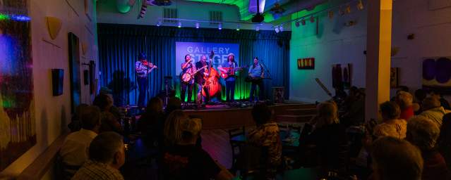 A bluegrass band made up of five members playing instruments such as a guitar, banjo, and bass, line a stage in an intimate music venue, playing before a crowd.