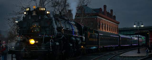 A steam engine decorated with Christmas lights and a holiday wreath on the engine is parked in front of a historic brick depot at night.
