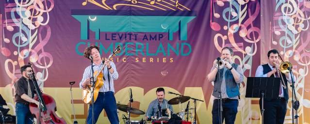 Full band on stage playing music in front of a colorful backdrop that says "Levitt AMP Cumberland Music Series."
