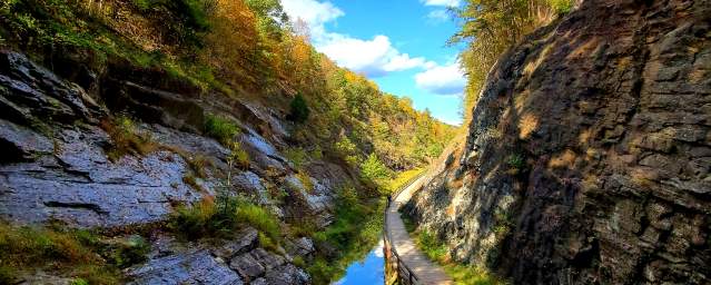 A scenic canal towpath runs alongside still water with dramatic rock walls and bright blue sky overhead.