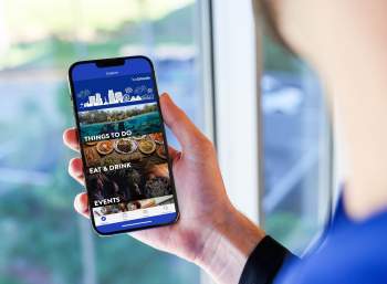 A person holding a smartphone displaying the VisitOrlando app interface, showcasing options like "Things to Do," "Eat & Drink," and "Events," with a blurred outdoor background.