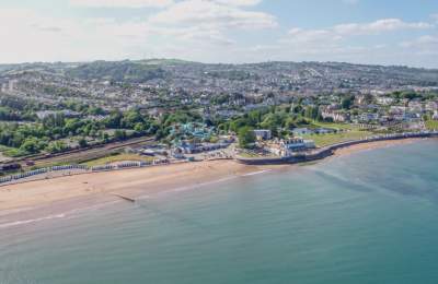 Aerial view of a coastal town with a sandy beach lined with blue beach huts. The sea is calm, and a scenic hillside town is in the background. The tone is peaceful.