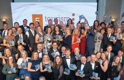 A large group of people dressed in formal attire are smiling and holding awards at an event. A screen in the background displays "tourism."