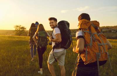 Four hikers with backpacks walk through a field at sunset. One glances back smiling, conveying a sense of adventure.
