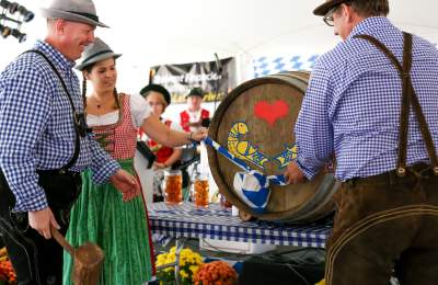 Attendees in traditional dirndl and lederhosen tap a decorated beer barrel at Oktoberfest in Lovettsville, Virginia.