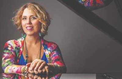 Gabriela Montero, a performer for the Waterford Concert Series, sits at a piano, wearing a colorful floral cardigan. The background is softly lit, enhancing her vibrant attire.