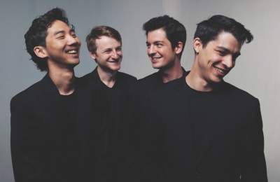 Quatuor Arod in black attire stand together, smiling and engaging with one another against a neutral background, creating a lively atmosphere.