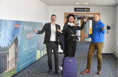 Three men posing in front of advertising boards for Shakespeare's England at Birmingham Airport. One is dressed in costume as William Shakespeare.