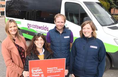 Four people smiling at the camera in front of a Go Cotswolds minibus holding up a red placard reading English Tourism Week